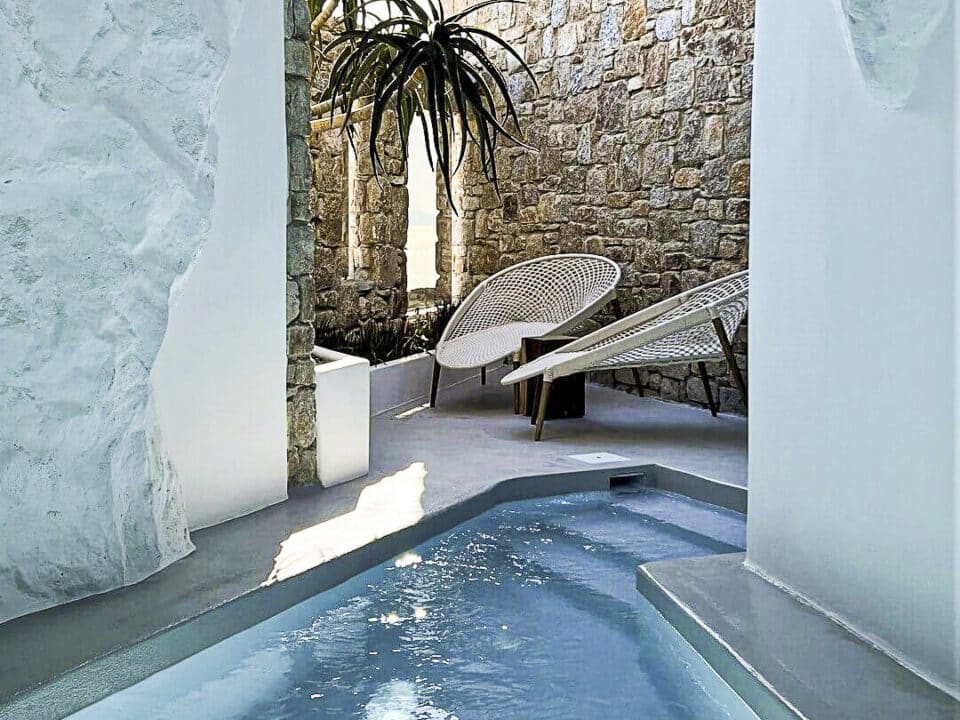 Nomia Mykonos Cave Pool Experience: Indoor pool with stone walls, lounge chairs, and sunlight reflecting on the water