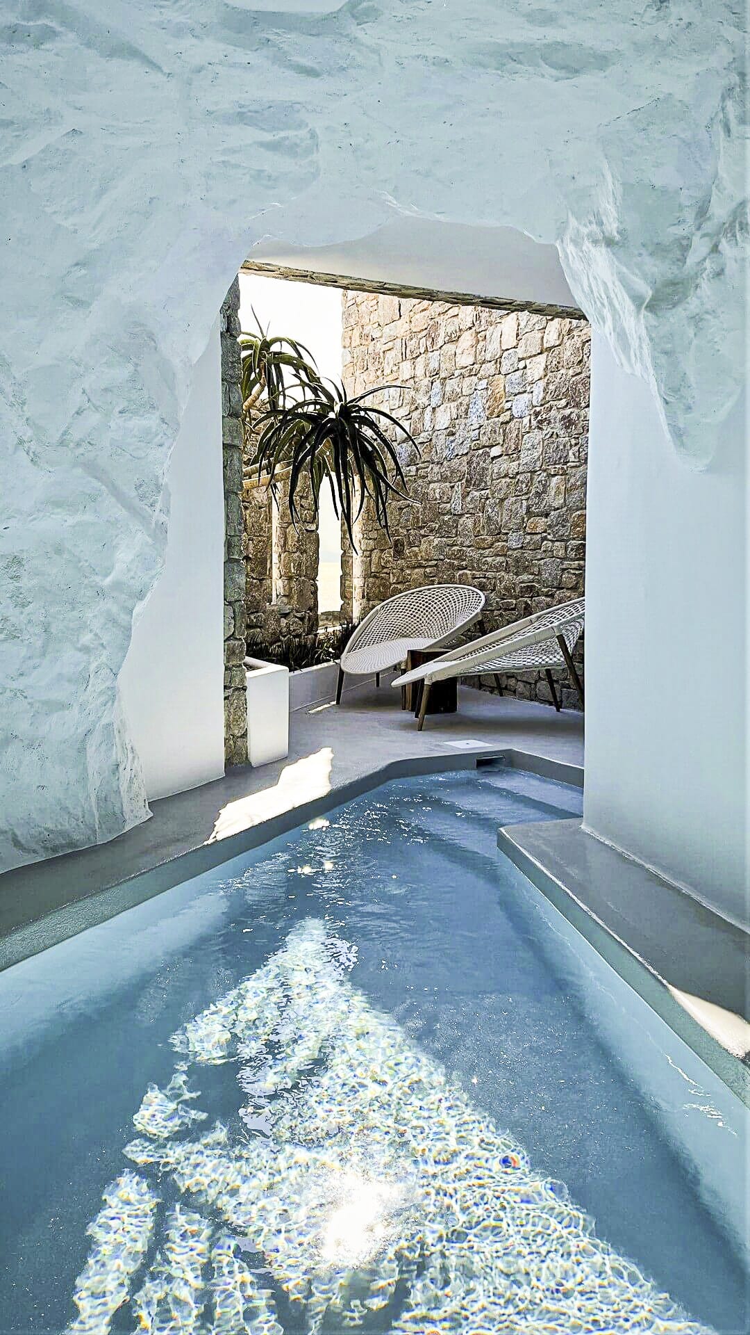 Nomia Mykonos Cave Pool Experience: Indoor pool with stone walls, lounge chairs, and sunlight reflecting on the water