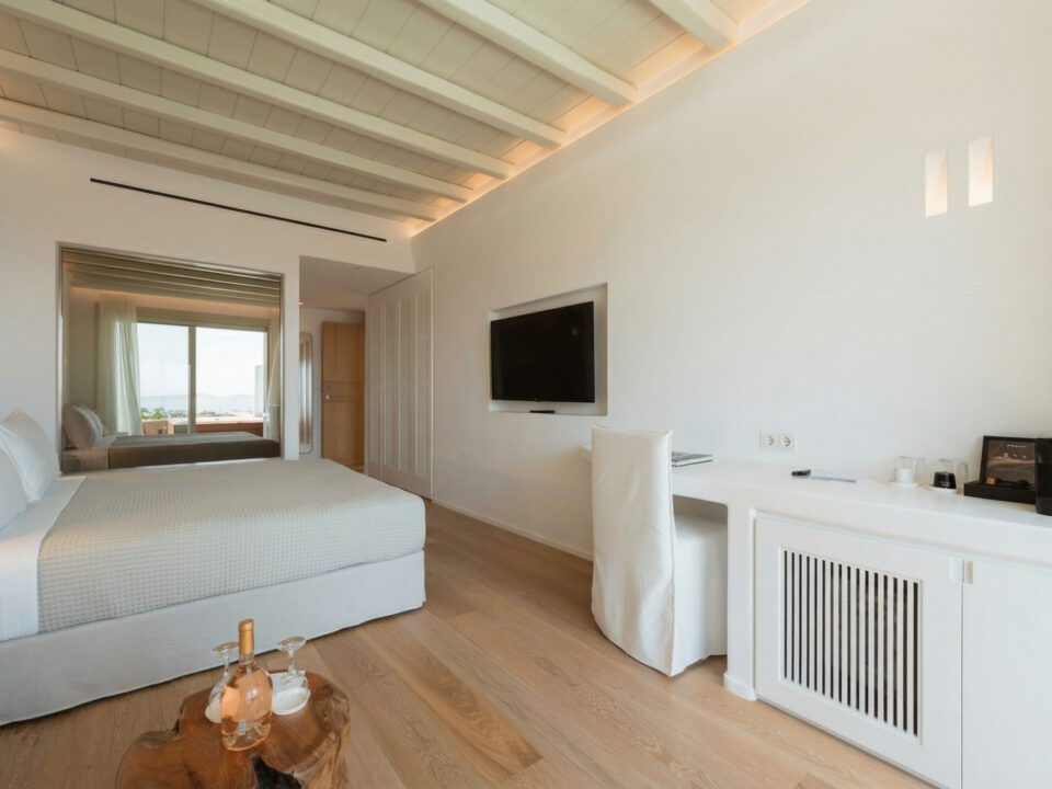 Deluxe Suite with Outdoor Hot Tub: Bright minimalist bedroom with a king-sized bed, wall-mounted TV, desk, and a view through the mirror to a balcony and sea beyond