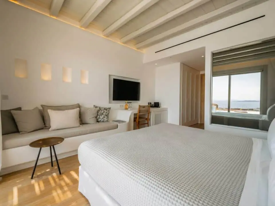 Junior Suite with Outdoor Hot Tub: Modern bedroom with a built-in sofa, TV, and a view of the sea reflecting on a large mirrored window
