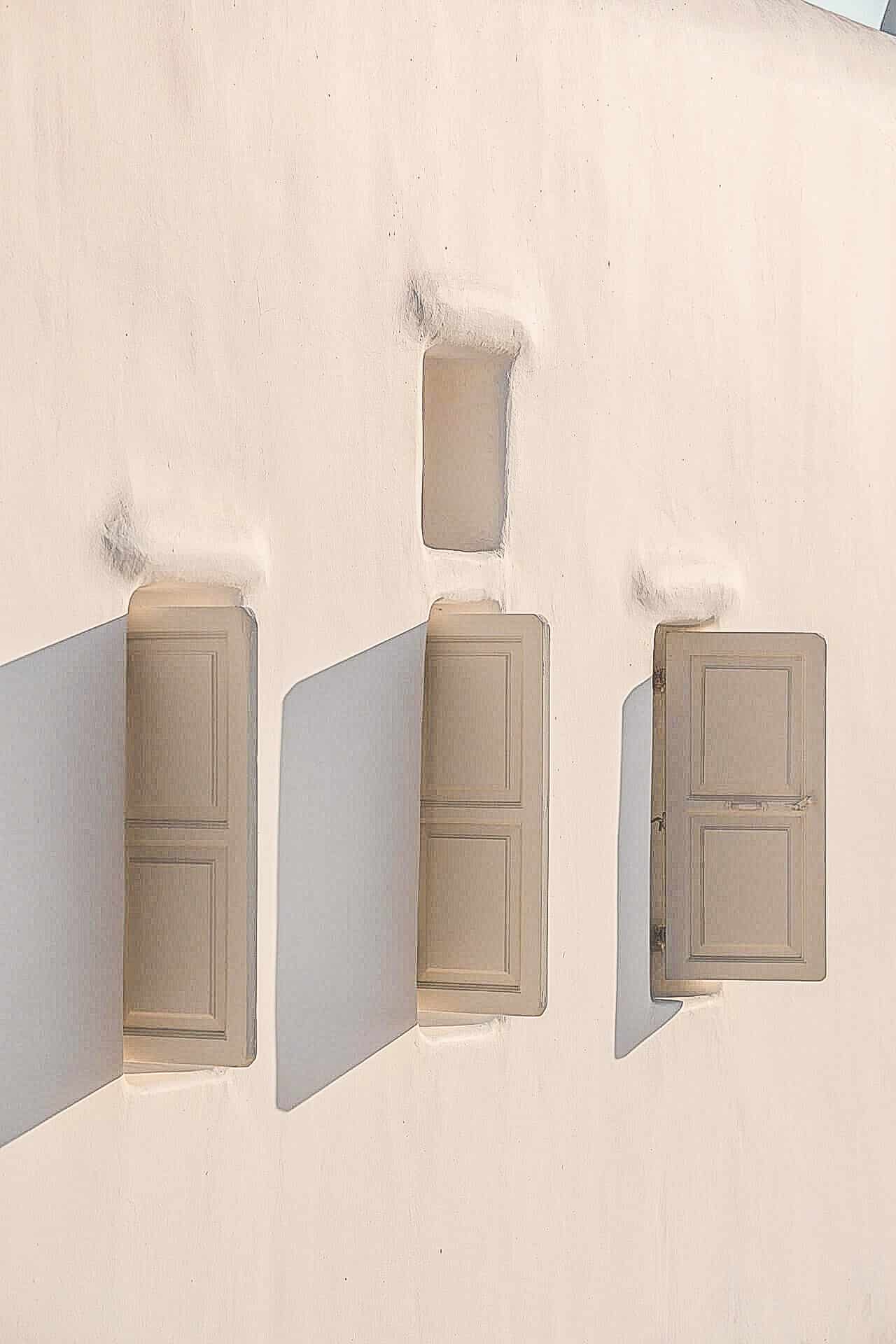 Nomia Mykonos Sunset Suites Architecture: Three beige wall-mounted shutters casting long shadows on a white wall