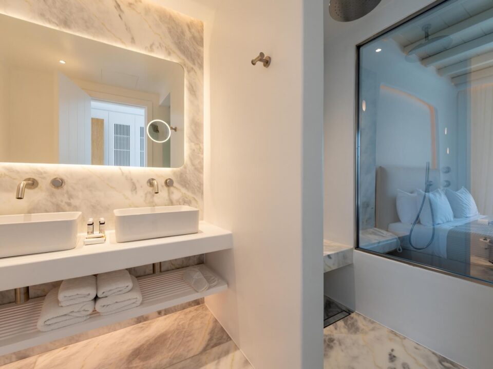 Junior Suite with Private Heated Swimming Pool: Luxury bathroom with double sinks, lit mirror, and a glass wall overlooking the bedroom