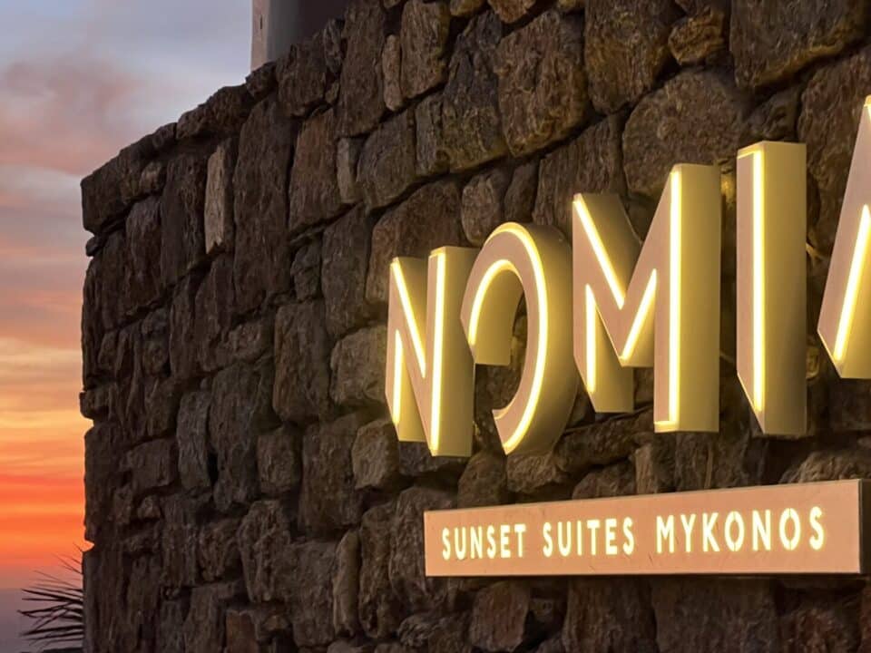 Illuminated NOMA sign on a stone wall at sunset, with “Sunset Suites Mykonos” below