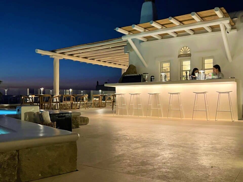 Nomia Mykonos Sunset Suites: Outdoor poolside bar at dusk with lit stools and two people behind the counter