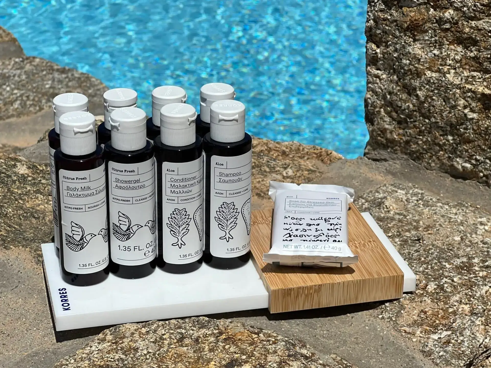 Nomia Mykonos Sunset Suites Amenities: Travel-size Korres toiletries on a tray by a pool, including body milk, shower gel, conditioner, shampoo, and soap